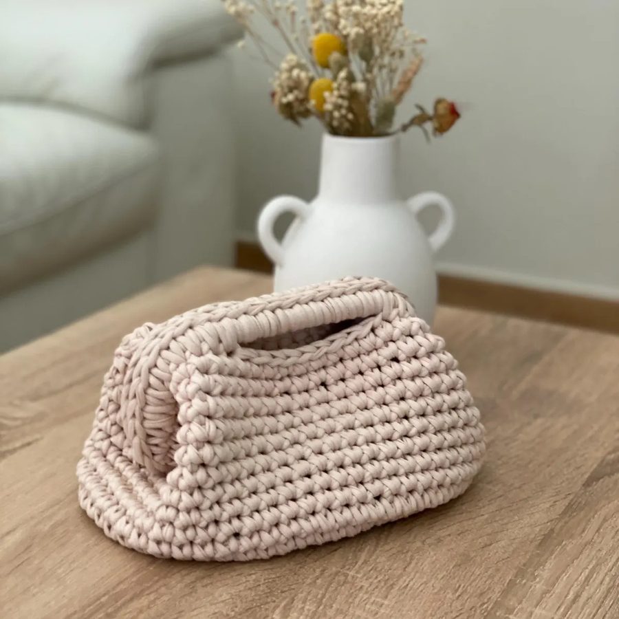 A blush T-shirt yarn clutch with a crocheted rim, on a wooden table beside a white vase of dried flowers.